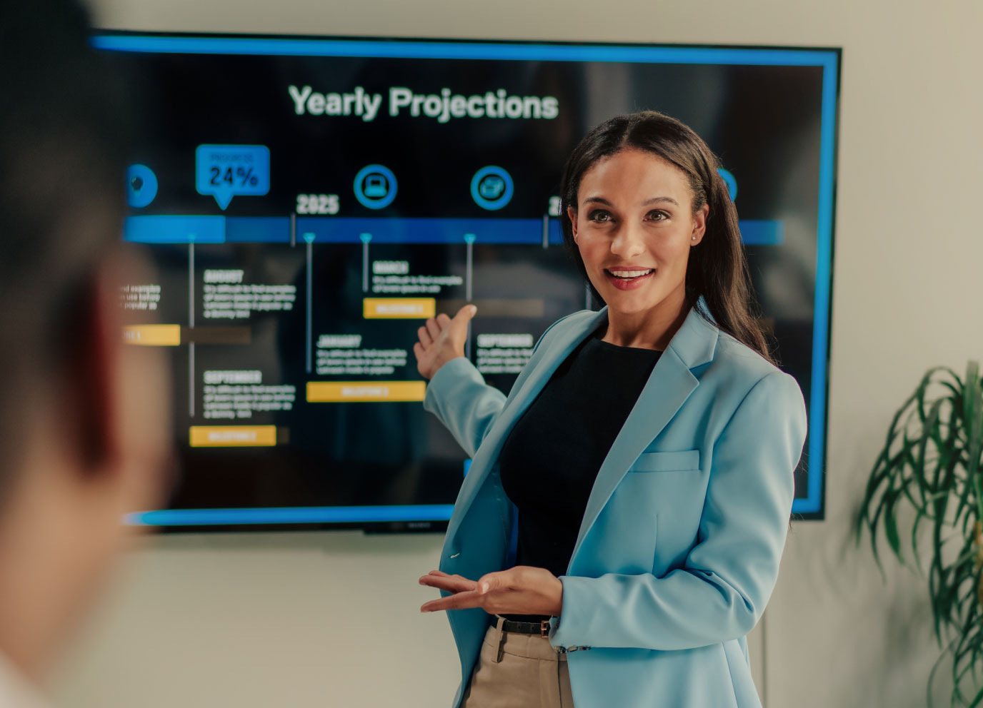 A woman dressed in business attire stands in front of a large presentation screen, engaging with the audience.