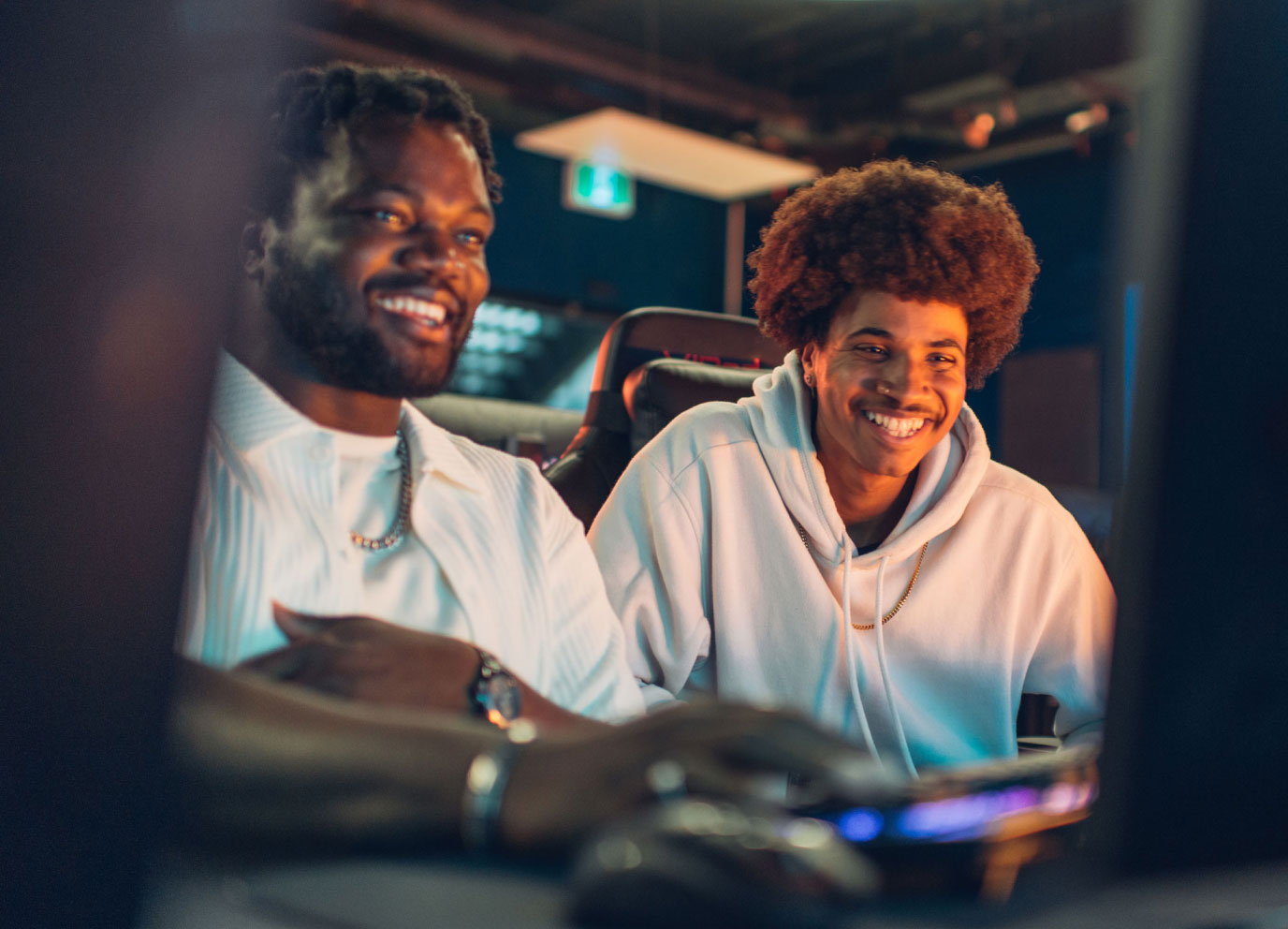Two students smiling while playing video games at a computer setup in a dimly lit room.